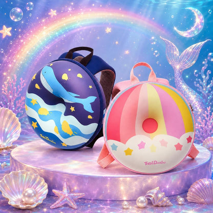 Two children's backpacks displayed on a glowing purple pedestal with a mermaid-themed fantasy background. One backpack is dark blue with a whale and star print, and the other is pink and yellow with a donut and star print.