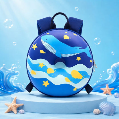 A round children's backpack with a whale and starry ocean design, displayed on a light blue pedestal with sea-themed props in a bright blue studio setting.